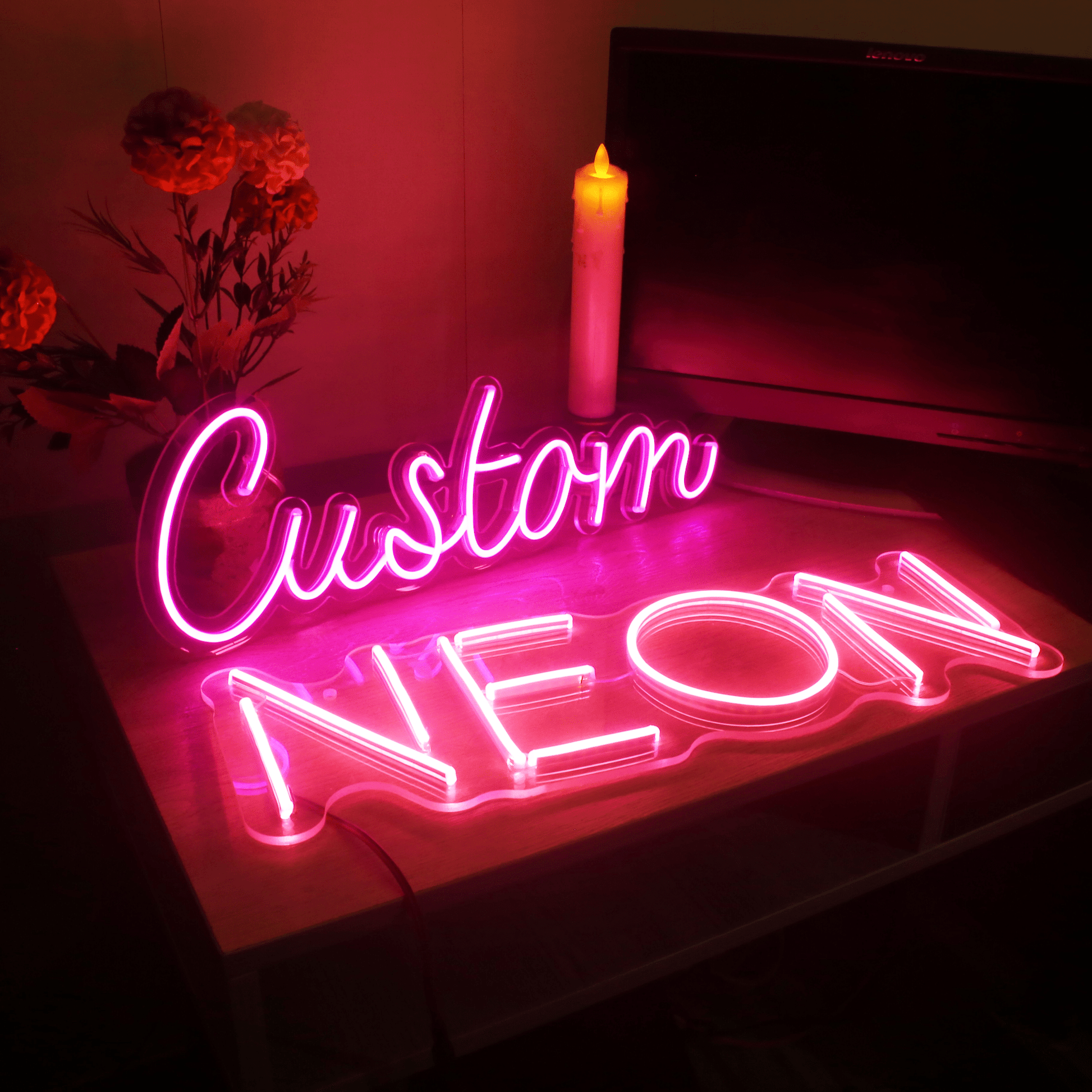 Pink bespoke neon sign spelling 'Custom NEON' displayed on a tabletop with soft ambient lighting, flowers and a candle in the background