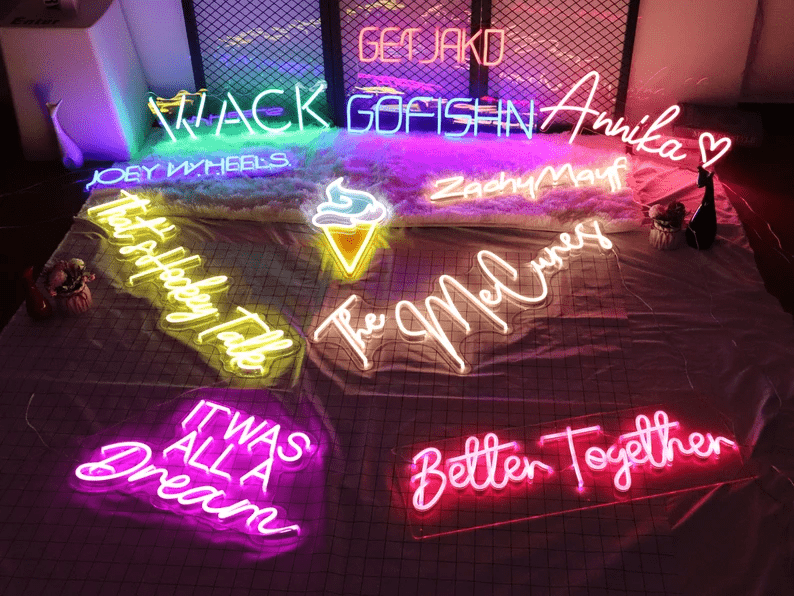 Assortment of custom neon signs laid out on fabric with phrases and names including 'Better Together', 'The McCunes', 'It Was All a Dream', 'Zachy Mayf', 'GOFISHIN', and an ice cream icon, in vibrant LED colours and unique fonts