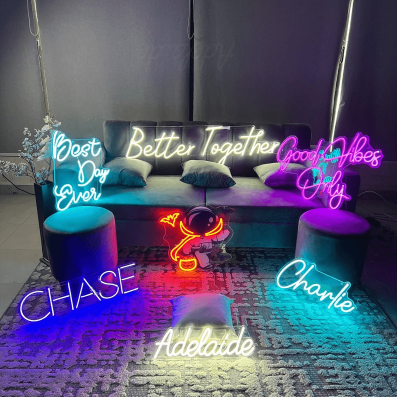 Collection of custom neon signs in various fonts and colours displayed in a modern lounge setup, featuring phrases like 'Better Together', 'Best Day Ever', 'Good Vibes Only' and personalised names including Chase, Charlie and Adelaide with a glowing astronaut design