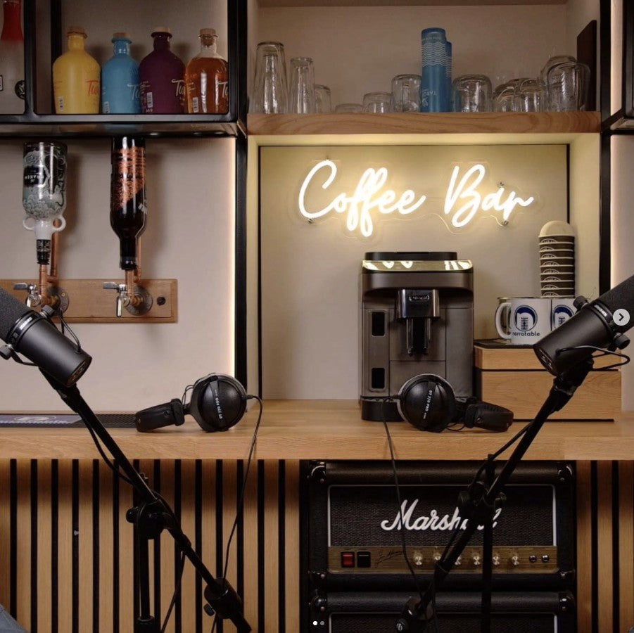 Coffee Bar LED neon sign in warm white backlit acrylic box above podcast studio desk with microphones and coffee machine backdrop