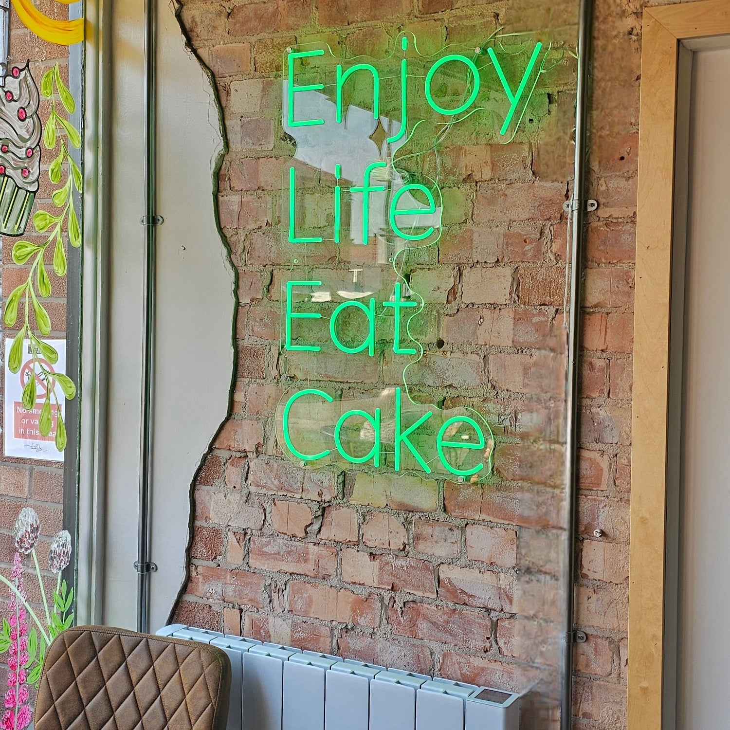 Green LED neon sign reading “Enjoy Life Eat Cake” mounted on an exposed brick wall inside a cozy café or cake shop