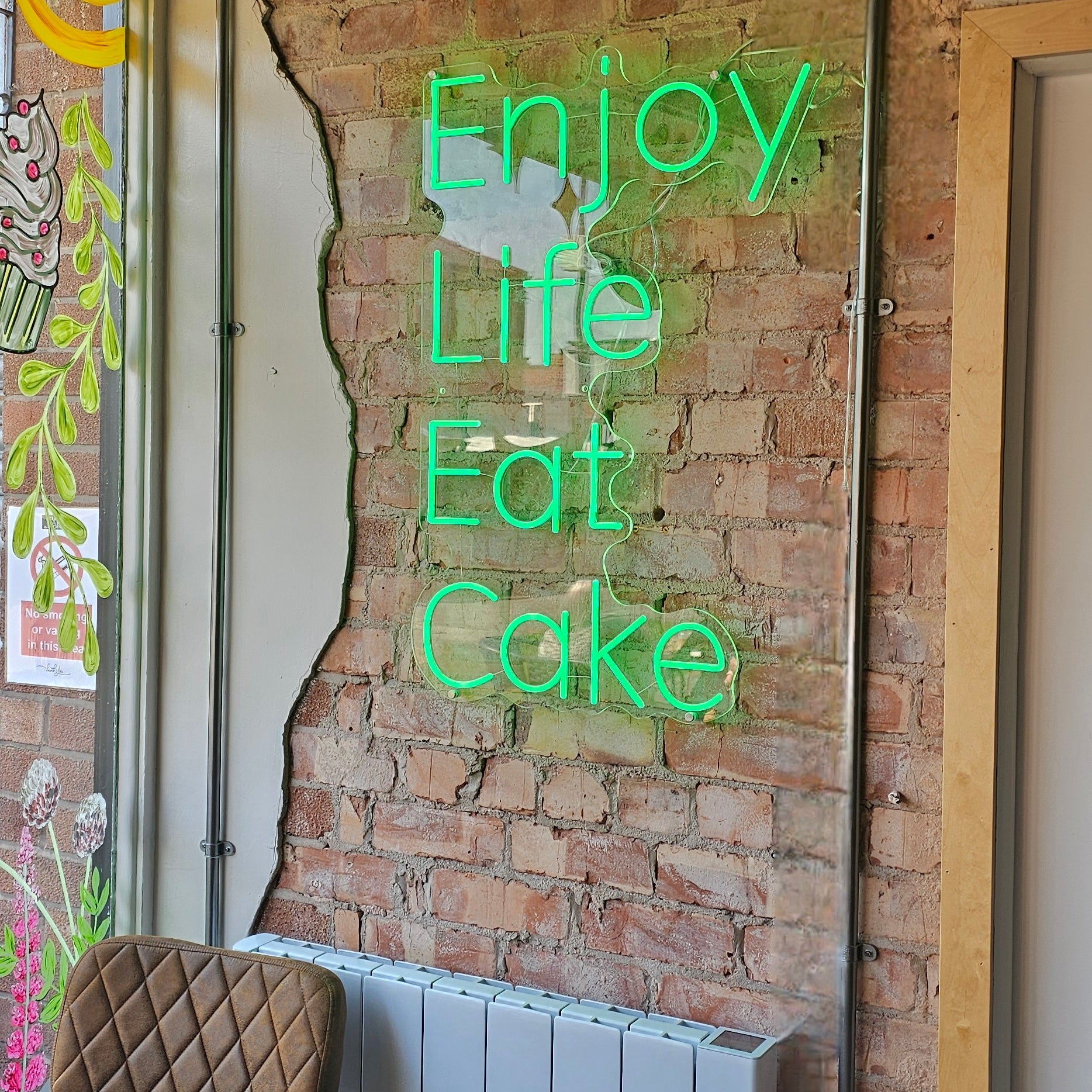 Green LED neon sign reading “Enjoy Life Eat Cake” mounted on an exposed brick wall inside a cozy café or cake shop