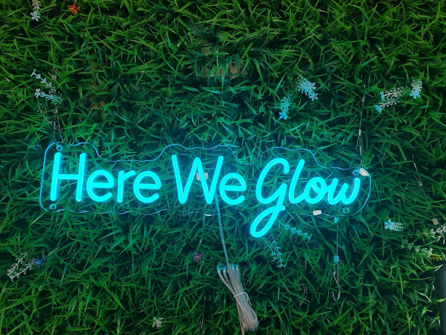 Cyan "Here We Glow" LED neon sign mounted on a green faux grass wall background for event decor.