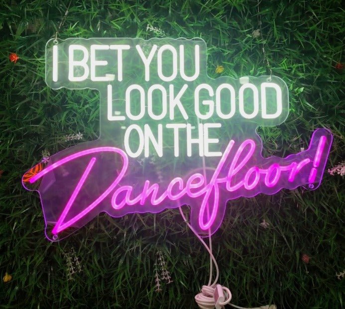 Dual-colour LED neon sign featuring "I Bet You Look Good On The" in white block letters and "Dancefloor!" in pink cursive, mounted on a green hedge wall.