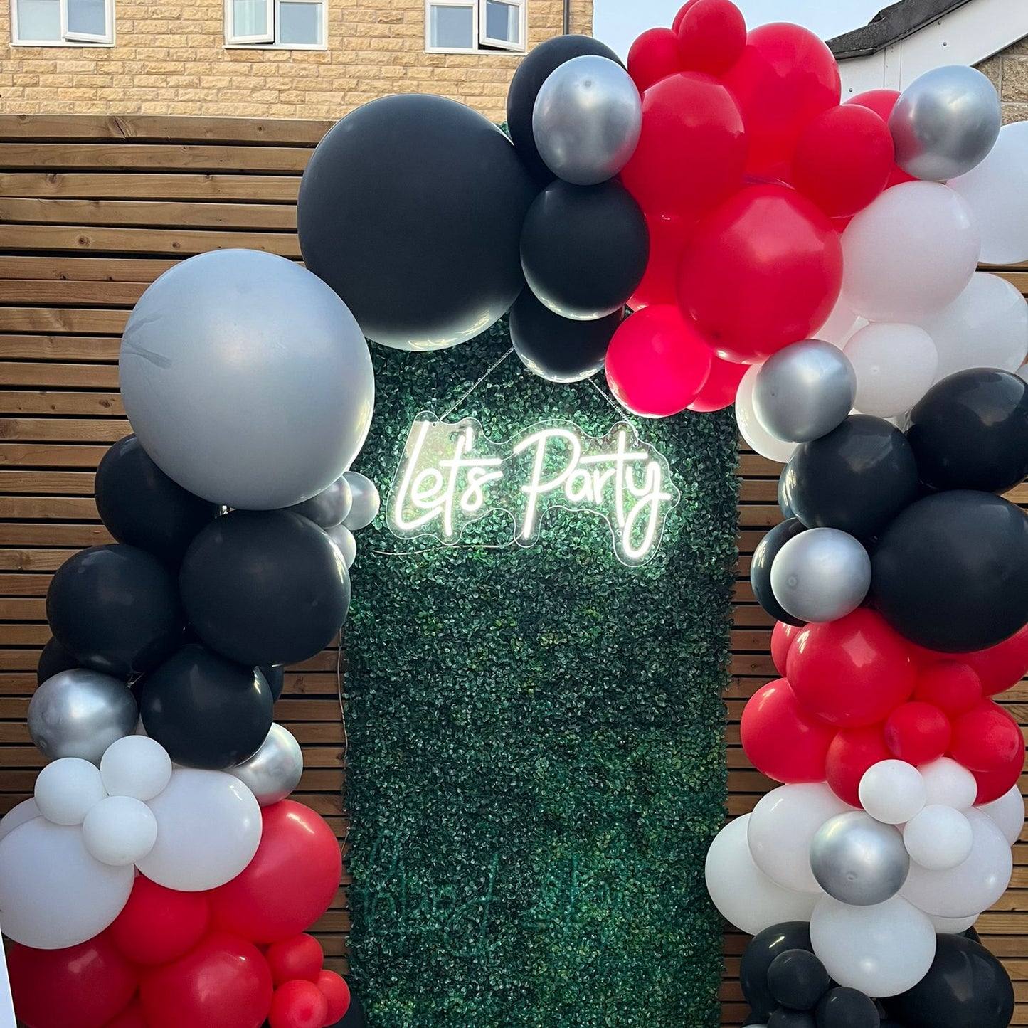 Let's Party LED neon sign on green wall backdrop with red, black, and white balloon arch — perfect for birthdays, events, and celebrations
