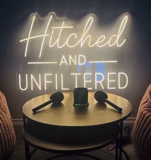 Warm white LED neon sign reading Hitched and Unfiltered in elegant script font behind podcast table with microphones, custom podcast studio signage UK