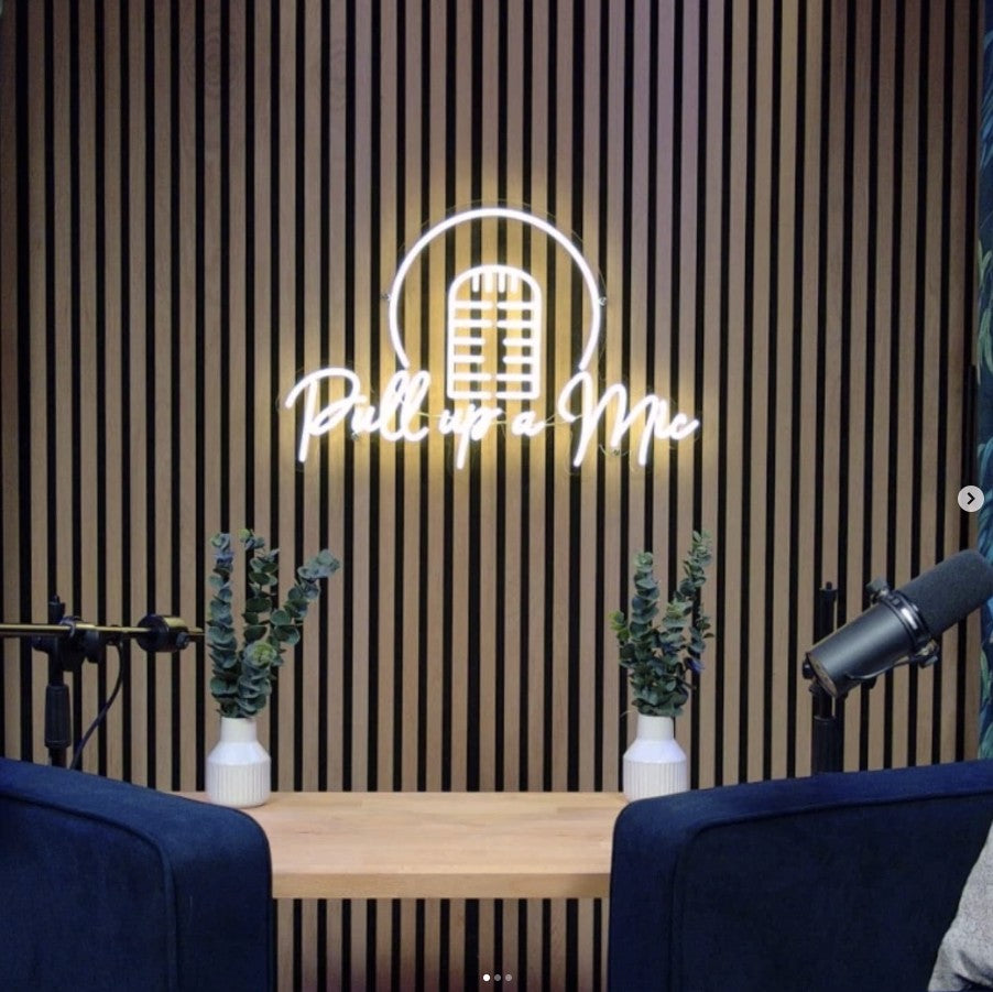 White LED neon sign reading Pull Up A Mic with microphone icon and headphone graphic on textured wooden slat wall podcast studio