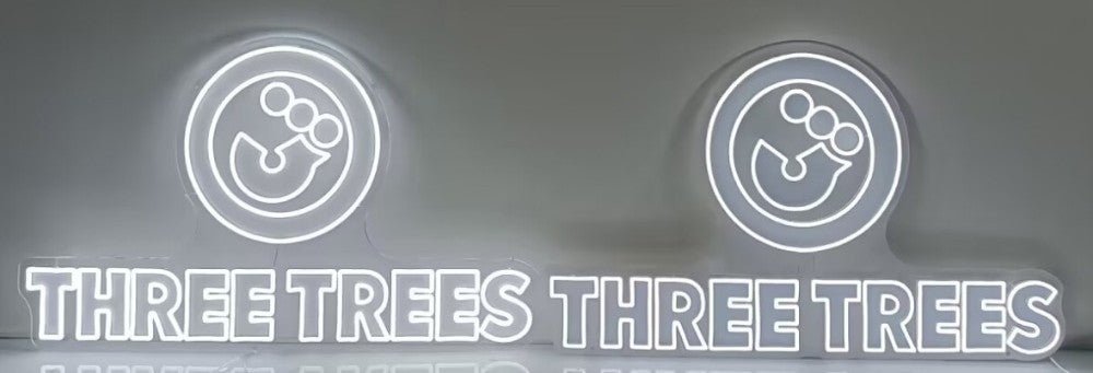 A side-by-side comparison of two illuminated white Three Trees LED neon signs. The split flexneon sign on the left is visibly brighter, whilst the integrated flexneon sign on the right displays a slightly dimmer, uniform line.