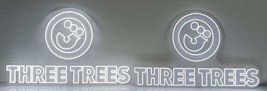 A side-by-side comparison of two illuminated white Three Trees LED neon signs. The split flexneon sign on the left is visibly brighter, whilst the integrated flexneon sign on the right displays a slightly dimmer, uniform line.