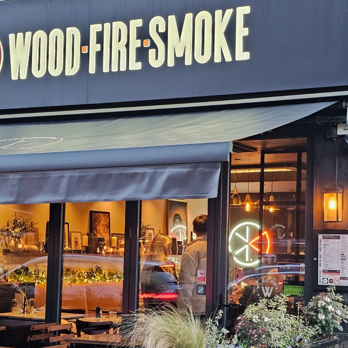 Custom restaurant logo LED neon sign designed for Wood Fire Smoke in Wilmslow, visible from the street inside the illuminated pizza restaurant window.