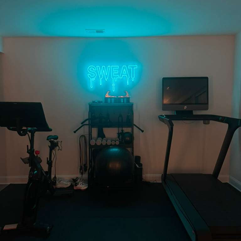 A glowing cyan blue LED neon sign with a dripping font effect that says 'SWEAT' mounted in a modern home gym between a treadmill and stationary bike.