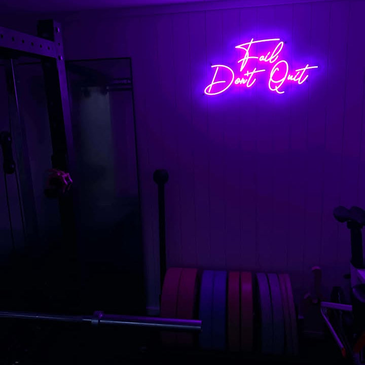 A vibrant purple LED neon sign displaying the motivational quote 'Fail Don't Quit' mounted on a wood-paneled wall in a dark home gym near weightlifting equipment.