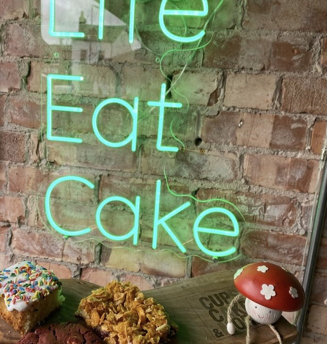 Green LED neon sign saying Life Eat Cake displayed in café with exposed brick wall and pastries, custom business signage UK