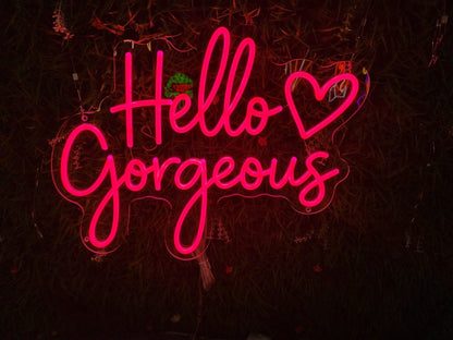 Hot pink "Hello Gorgeous" cursive LED neon sign with a heart icon, mounted on a dark foliage wall for beauty salon or bedroom decor.