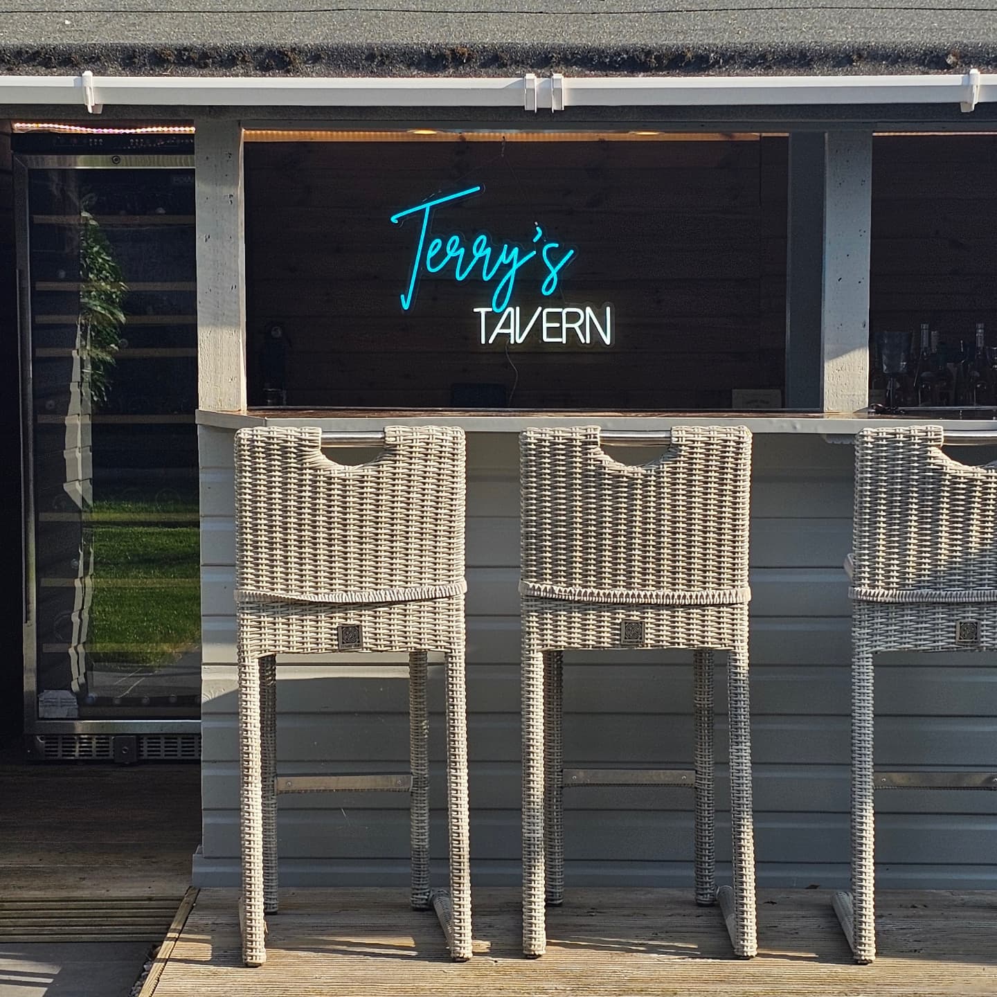 Custom home bar LED neon sign reading Terry's Tavern in blue and white script mounted on outdoor garden bar with bar stools UK