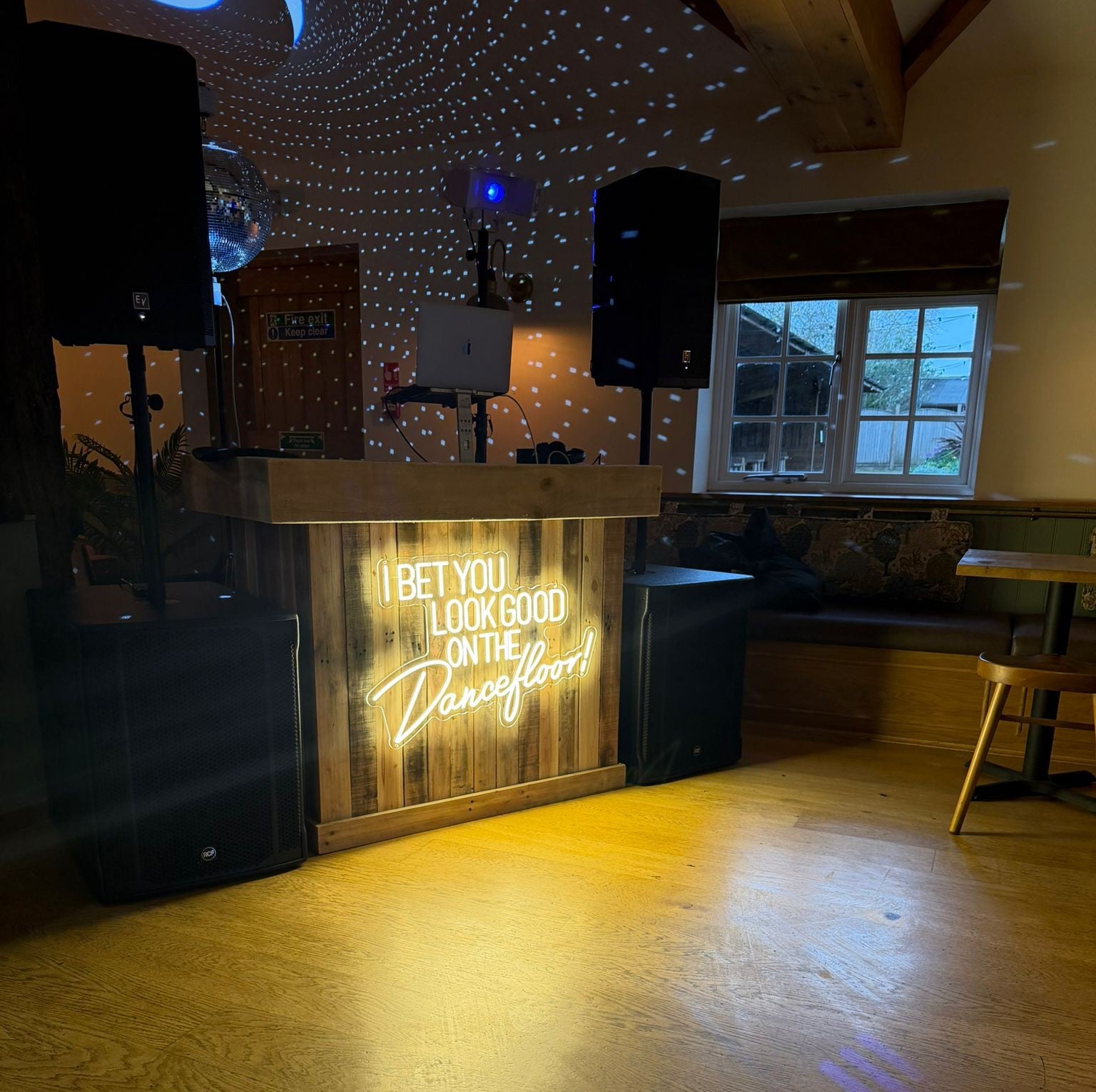 Warm white "I Bet You Look Good On The Dancefloor" LED neon sign mounted on a rustic wooden DJ booth with disco ball lighting and party speakers.