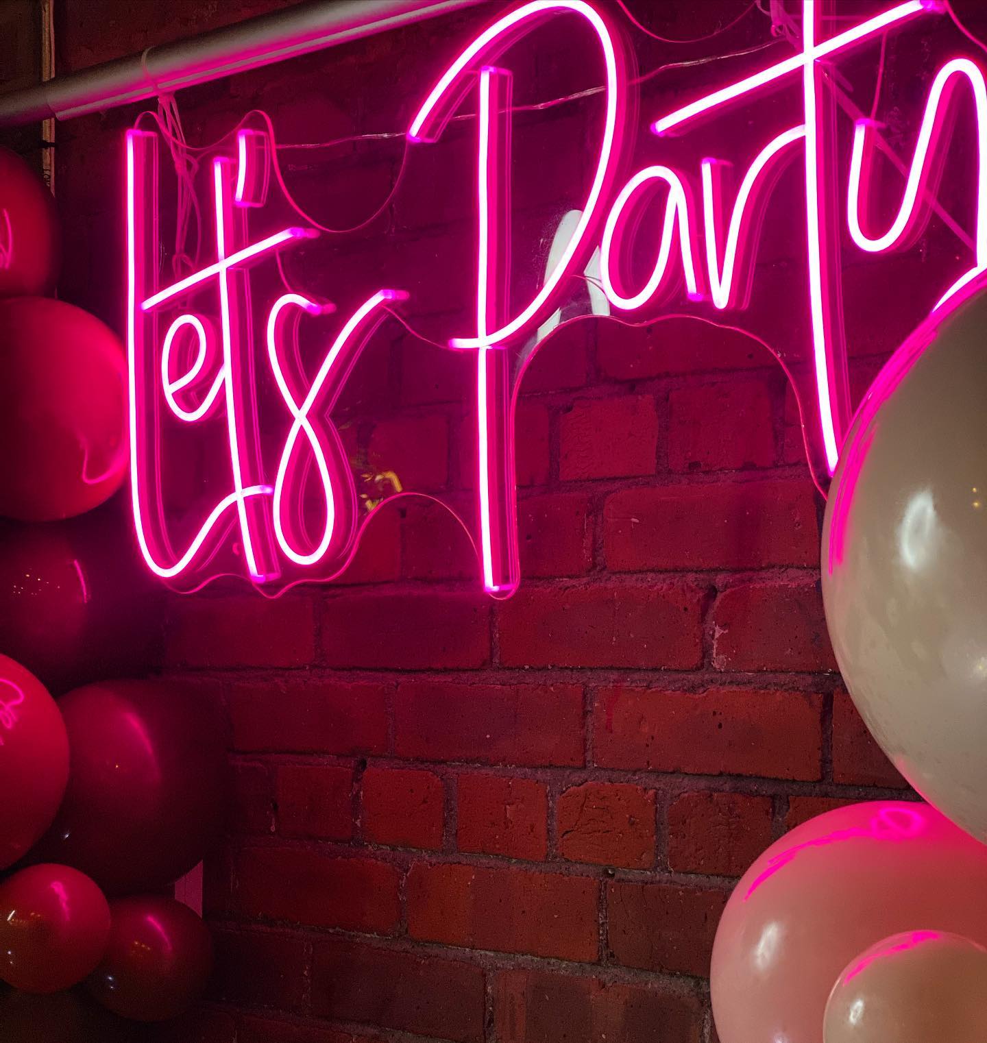 Close-up of a pink "Let's Party" neon sign hanging on a rustic red brick wall surrounded by pink and white party balloons, ideal for birthday or engagement decor.