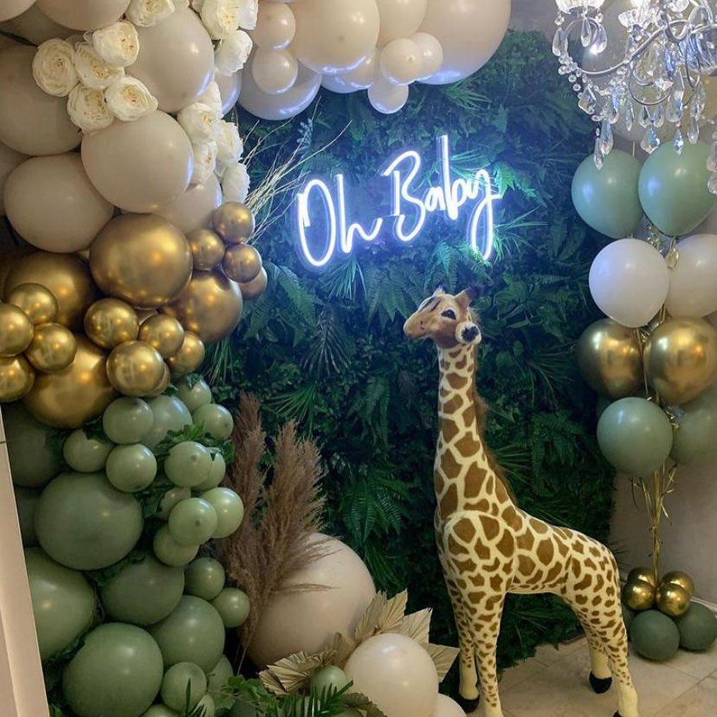 Modern white LED "Oh Baby" neon sign centrepiece for a jungle-themed baby shower, featuring a large stuffed giraffe, gold balloons, and tropical greenery.