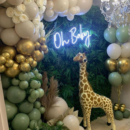 Modern white LED "Oh Baby" neon sign centrepiece for a jungle-themed baby shower, featuring a large stuffed giraffe, gold balloons, and tropical greenery.