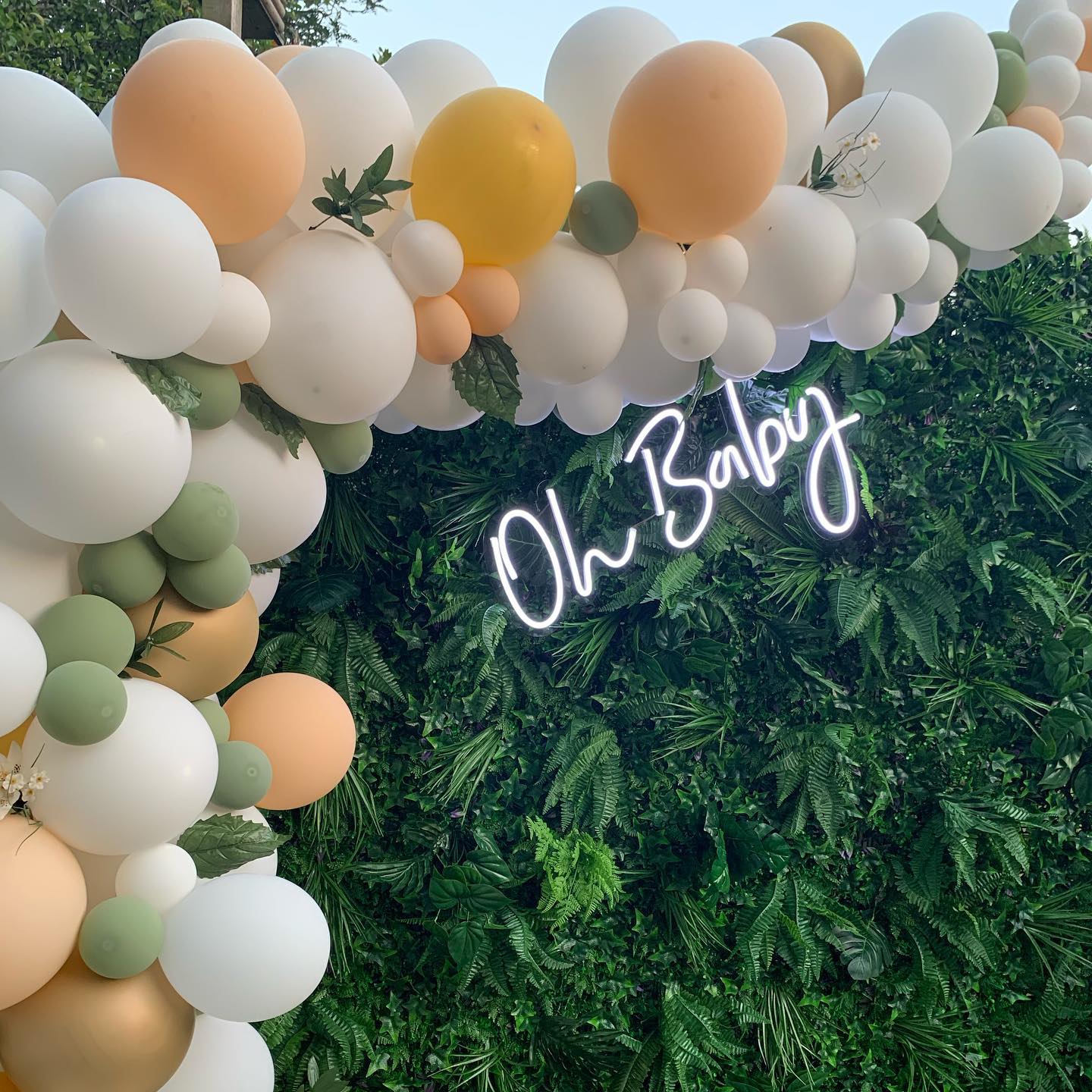 A glowing white "Oh Baby" neon sign mounted on a lush green fern and ivy foliage backdrop, framed by a soft-toned balloon arch in sage green, peach, and white.