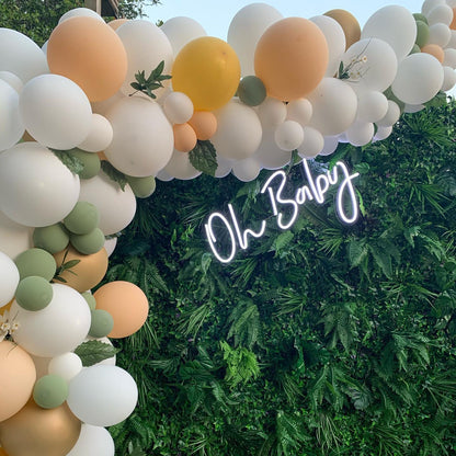 A glowing white "Oh Baby" neon sign mounted on a lush green fern and ivy foliage backdrop, framed by a soft-toned balloon arch in sage green, peach, and white.