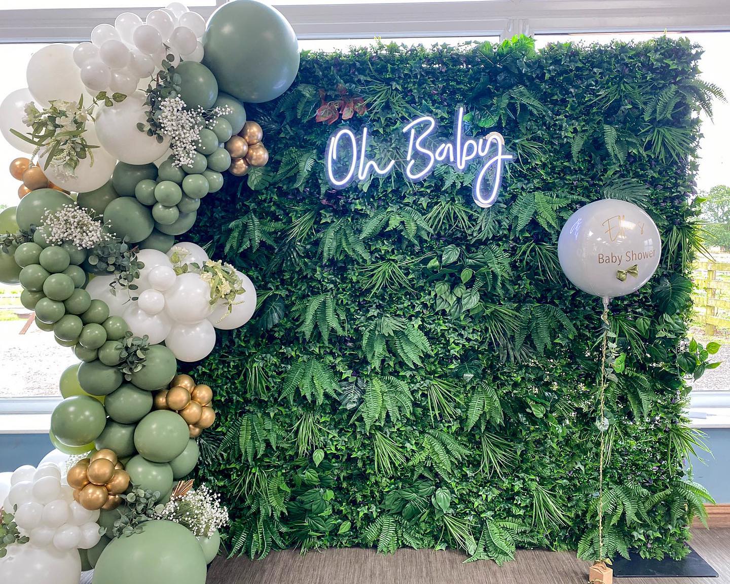 Cold white "Oh Baby" neon sign hanging on a dense green hedge wall, framed by a sage green, peach, and white balloon garland for a gender-neutral baby shower.