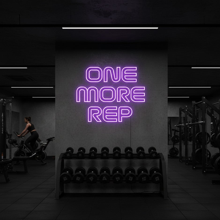 A large purple LED neon sign saying 'ONE MORE REP' mounted on a concrete pillar in a modern commercial gym with black rubber mats, hexagonal dumbbells on a rack, and black strength training machines.