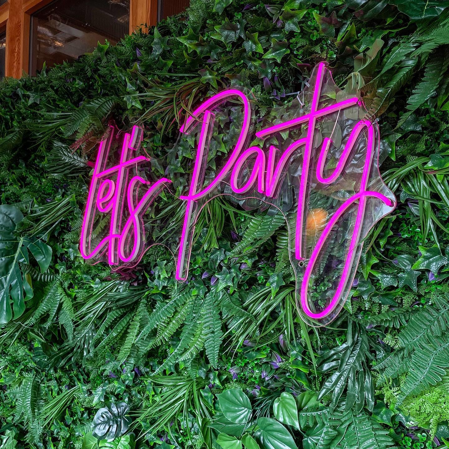Hot pink "Let's Party" LED neon sign mounted on a lush green faux foliage grass wall, creating a vibrant photo backdrop for events and parties.