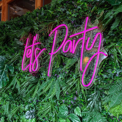 Hot pink "Let's Party" LED neon sign mounted on a lush green faux foliage grass wall, creating a vibrant photo backdrop for events and parties.