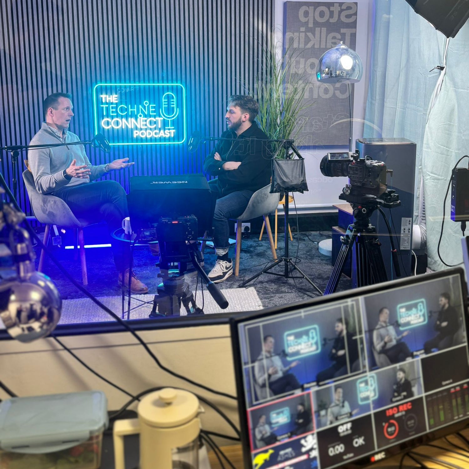 Custom podcast neon sign for The Techie Connect Podcast glowing blue in a recording studio during filming, with hosts and cameras in shot
