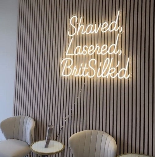 Warm white LED neon sign reading Shaved Lasered BritSilk'd in elegant script font for beauty salon on textured wooden wall backdrop